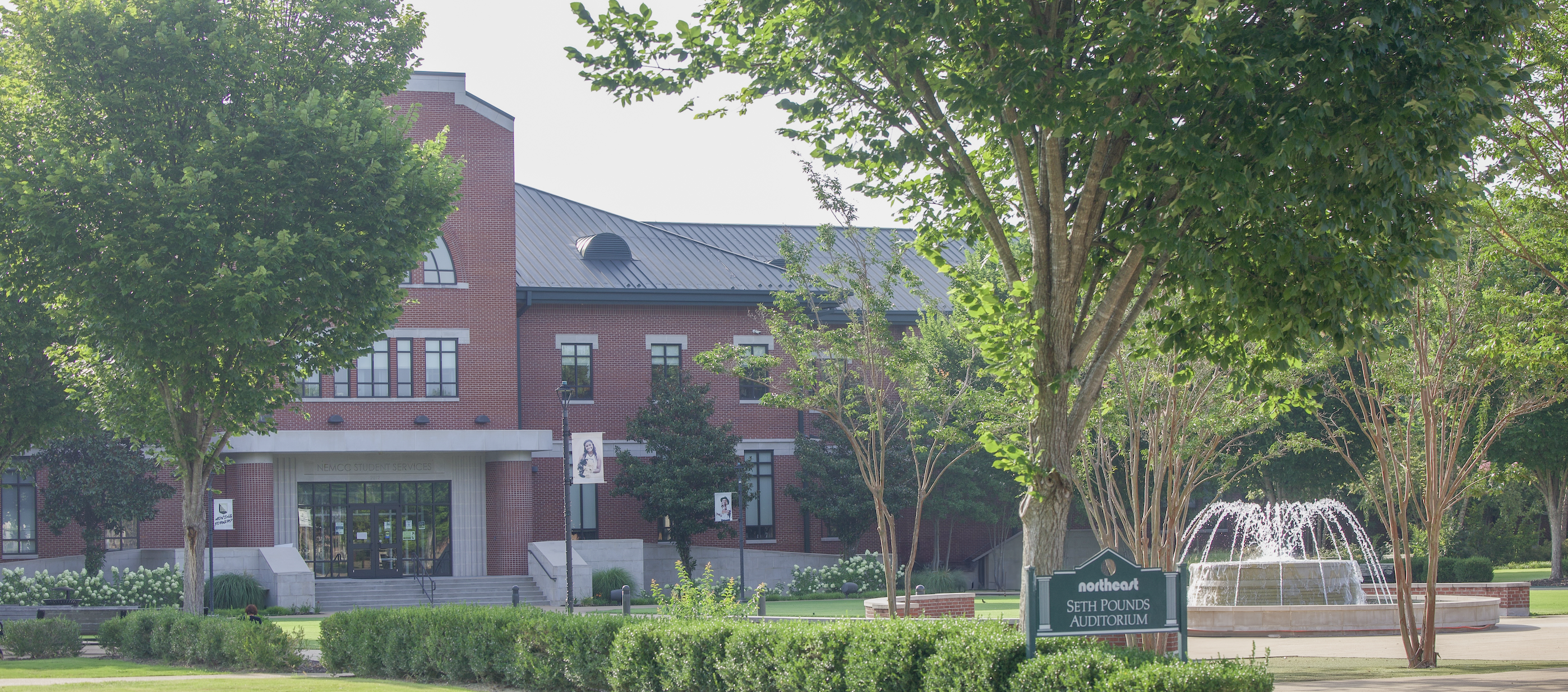 Brick campus building framed by leafy trees, with a fountain and a sign for Seth Ponds Auditorium in the foreground.