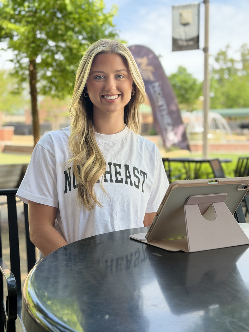 female student with blonde hair wearing a white Northeast tshirt.  She is sitting at a table with an iPad smiling at the camera.