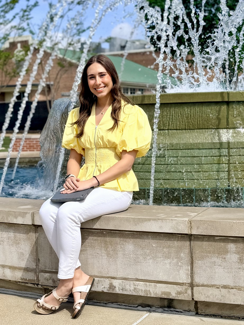 female in a yellow shirt and white jeans sitting in front of a fountain with an iPad in her lap