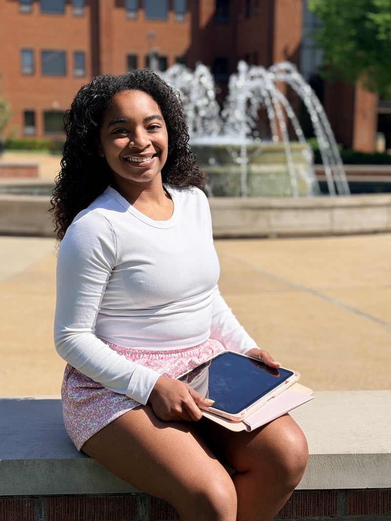 female student wearing a white shirt and pink shorts sitting in front of a fountain holding an iPad